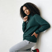 Woman wearing a green sweatshirt and gray pants against a white wall