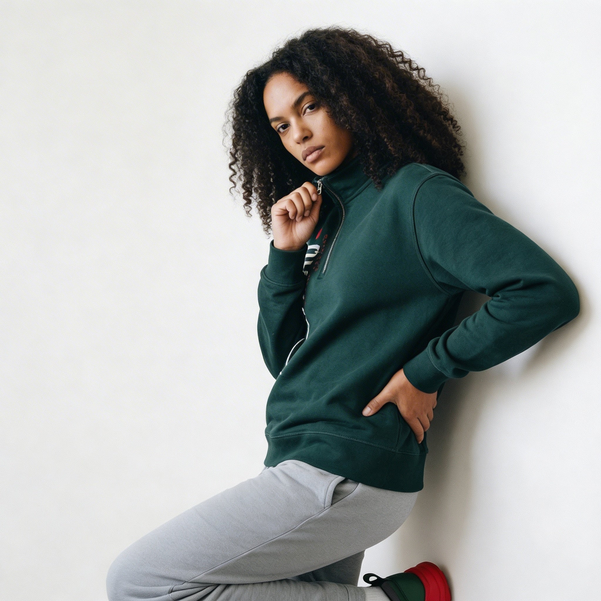 Woman wearing a green sweatshirt and gray pants against a white wall