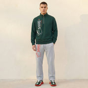 Man wearing a green sweatshirt and gray pants with branding, standing against a plain background