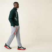 Man wearing a green jacket and gray pants with brand logos, walking on a beige background