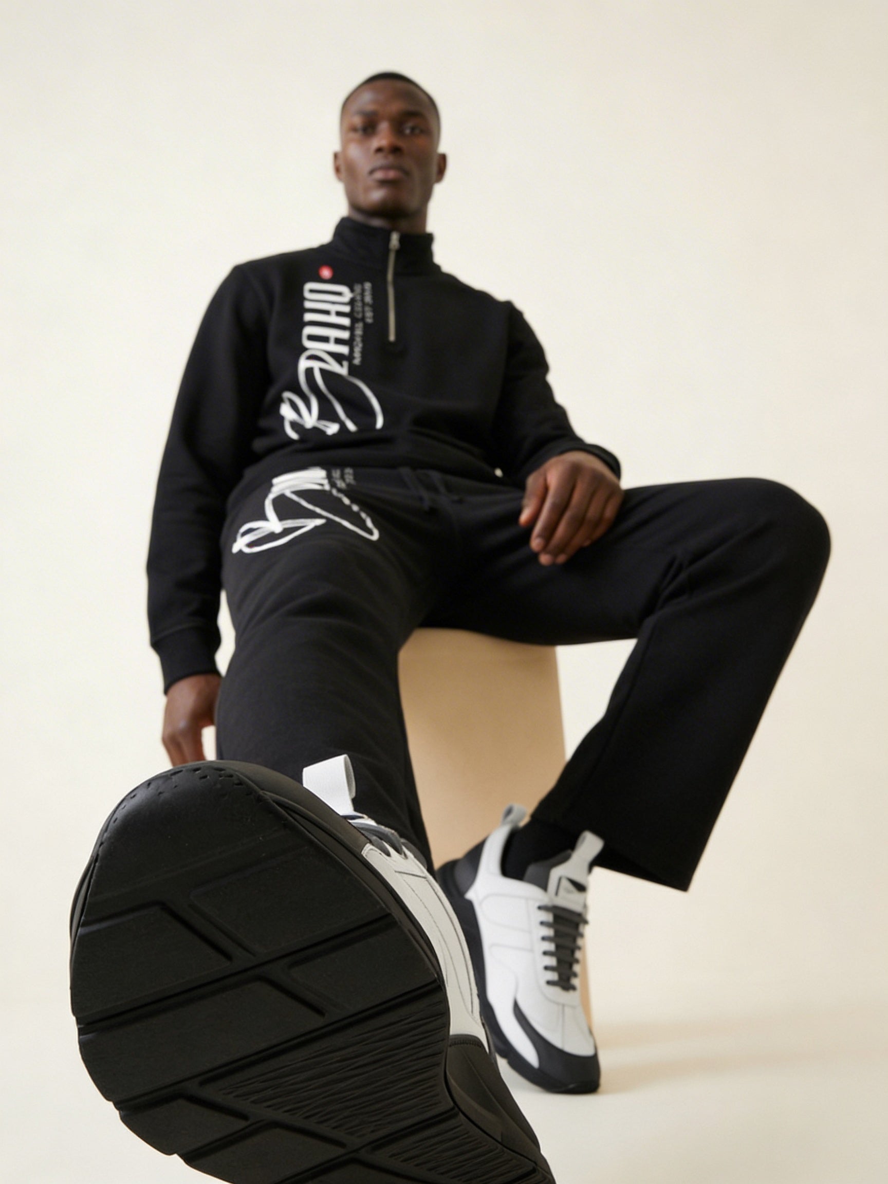 Man wearing a black tracksuit with white text, sitting on a beige stool.