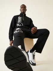 Man wearing a black tracksuit with white text, sitting on a beige stool.