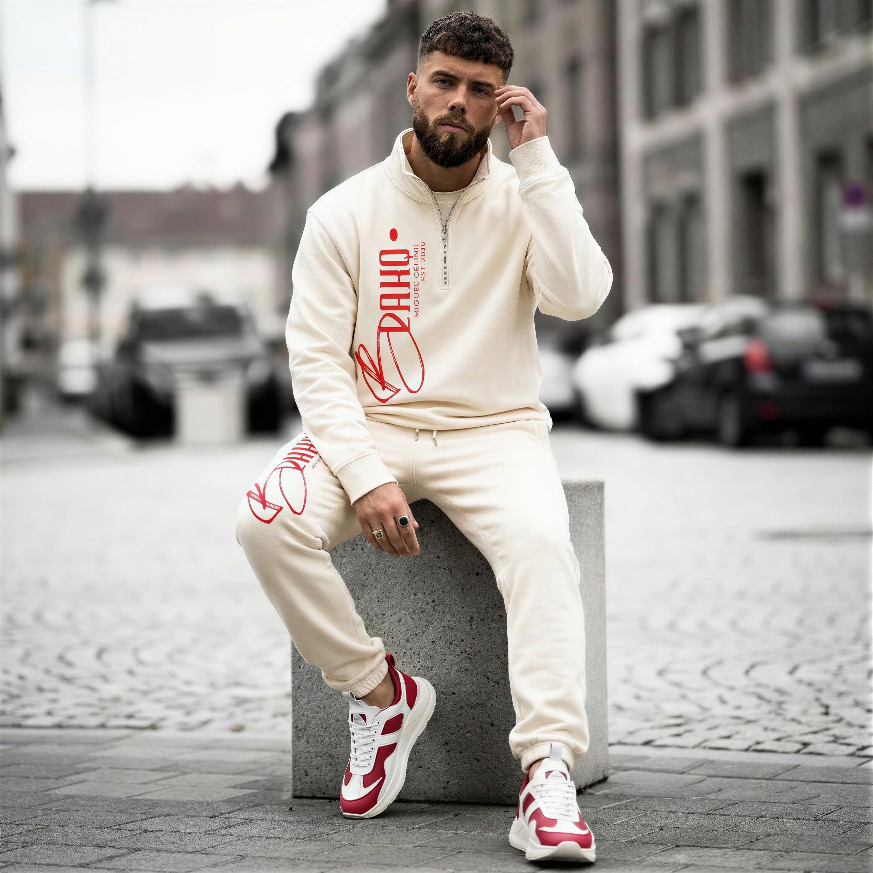 Man wearing a cream tracksuit with red branding sitting on a concrete block in an urban setting.