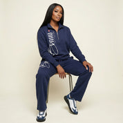 Woman wearing a navy tracksuit with white text sitting on a chair against a beige background