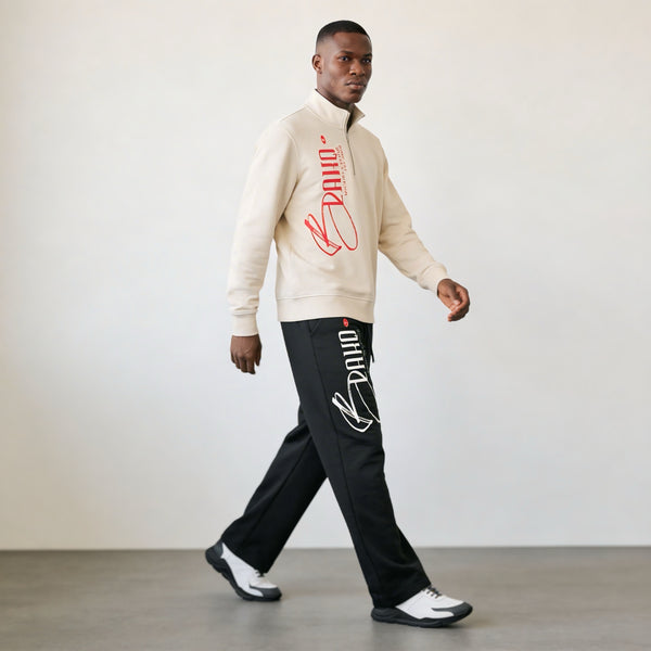 Man wearing a beige jacket and black pants with brand logos, walking against a plain background